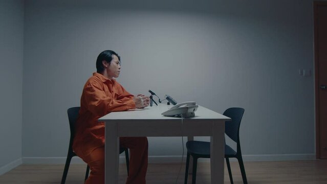 Arrested Asian man in orange jumpsuit and handcuffs sitting at table in interrogation room, waiting for an interview. Zoom Shot