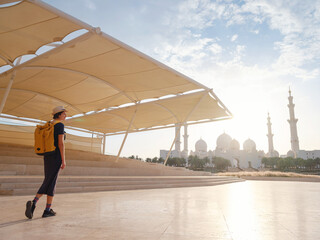 travel to the United Arab Emirates, Happy young asian female traveler with backpack and hat in Wahat Al Karama or Oasis of Dignity, permanent memorial for its martyrs, and Shaikh Zayed Grand Mosque.