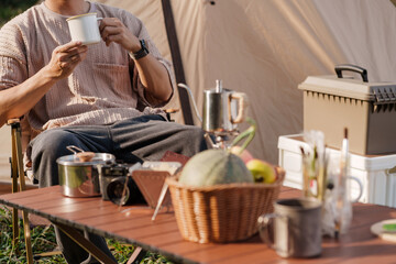 Fototapeta na wymiar Asian man drinking coffee enjoying camping outdoors in nature. Man traveler hands holding cup of coffee.
