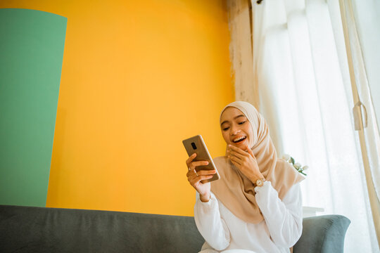 portrait of Asian veiled woman laughing sitting on sofa using cell phone at home