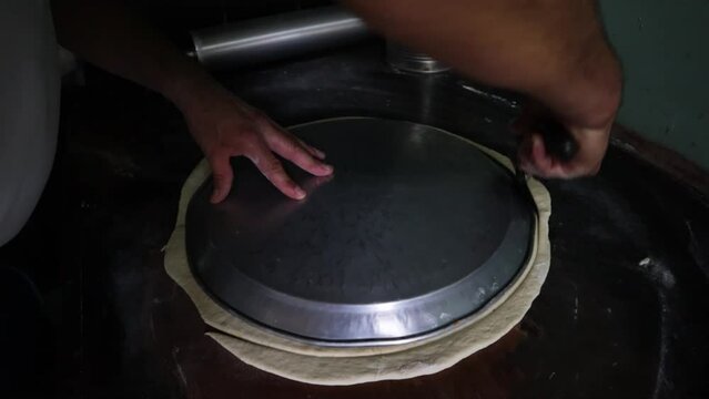 A slow motion handheld shot of a man cutting dough in a circular shape with a pizza cutter and a metal tray to prepare a pizza in a restaurant kitchen.