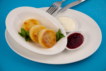 Round cottage cheese pancakes in a white plate on a blue background. Food breakfast. Healthy food. Sour cream sauce and mint. Top view closeup. Dessert meal. Curd cheese pancake ricotta fritters.