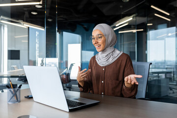 Successful and happy woman inside office at workplace, businesswoman boss in hijab talking on video call using laptop sitting at table muslim woman joyfully gesturing to colleagues in online meeting - obrazy, fototapety, plakaty