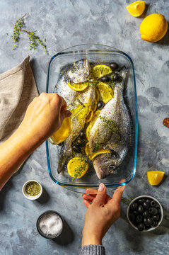 Two raw fresh sea bream or dorada are ready for cooking with lemon and spices in a glass pot, woman's hand pouring oil to the dish. Grey concrete background, napkin, top view.