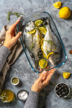 Two raw fresh sea bream or dorada are ready for cooking with lemon and spices in a glass pot, the woman's hands holding the dish. Grey concrete background, napkin, top view.