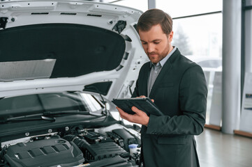 Standing, side view, holding tablet. Man in black suit is indoors in the car dealership near automobile