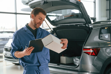 With box and notepad. Man in blue uniform is working in the car service