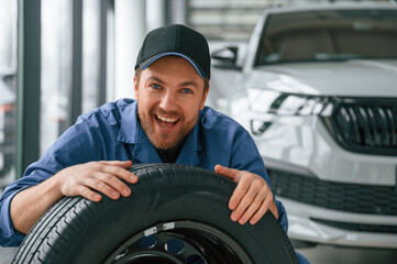 Fototapeta na wymiar New tire. Leaning on it. Man in blue uniform is working in the car service