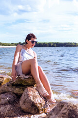 Winsome Relaxed Sensual Caucasian Brunette Female Girl in Casual Summer Beach Clothing Sitting On Stone on Coastal or Sea Shoreline