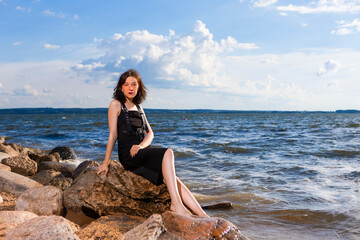 Winsome Relaxing Caucasian Brunette Girl Posing in Black Dress On Stone Line At Sea During Sunny Day Outdoors.