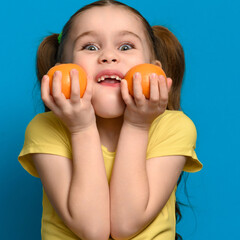 Fototapeta na wymiar A small and smiling girl is holding two tangerines on a blue background.