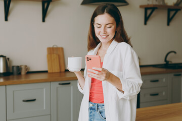 Young smiling happy fun cheerful housewife woman wearing casual clothes drink coffee use mobile cell phone chat online eat breakfast cooking food in light kitchen at home alone. Healthy diet concept.