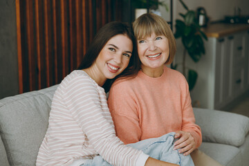 Obraz premium Two adult smiling fun women mature mom young kid wearing casual clothes look aside posing hug sit on gray sofa couch stay at home flat rest relax spend free spare time in living room. Family concept.