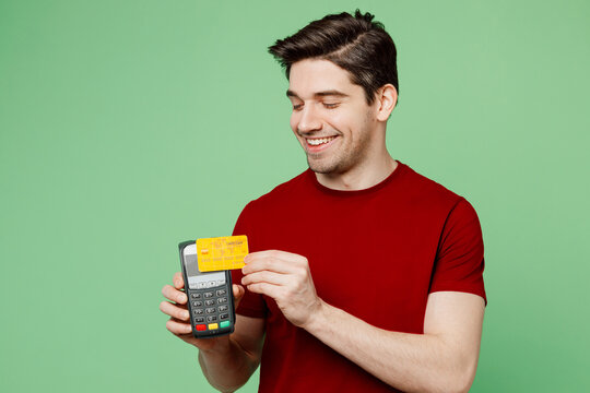 Young man wears red t-shirt casual clothes hold wireless modern bank payment terminal to process acquire credit card isolated on plain pastel light green background studio portrait. Lifestyle concept.