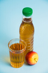 Apple juice drink in plastic bottle and reusable glass cup on blue paper background. Concept of earth day, zero waste and plastic recycling.