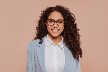 Obraz premium Face portrait of a young multiracial beautiful happy smiling businesswoman with curly hair, wears blue suit and eyeglasses, isolated over beige background at studio. Business concept.