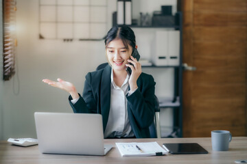 Portrait of Asian businesswoman  talking mobile phone and working on laptop and papers on table in her office.