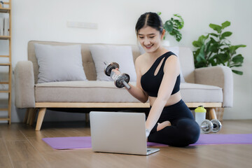 Sporty woman in sportswear is sitting on the floor with dumbbells and a protein shake or a bottle of water and is using a laptop at home in the living room.