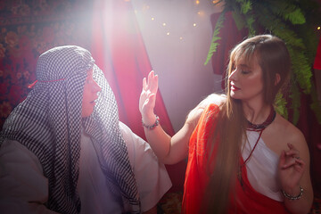 Portrait of young arabian muslim couple in traditional clothes in cozy red room. Fhoto shoot in easten style with male and female model like in a harem with a sultan and an odalisque. Partial focus