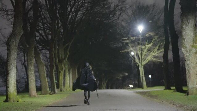 A young female child dressed for Halloween as a plague Doctor walks briskly in between trees at night