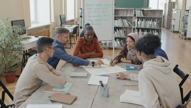 Teacher of English language and ethnically diverse migrant students sitting around table making educational poster together