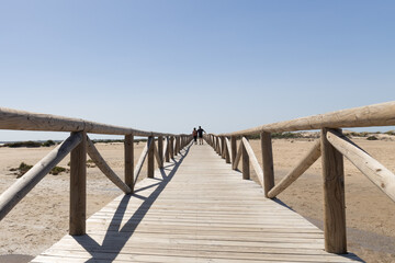 Una pareja paseando sobre una pasarela de madera en una playa del sur de Andalucía en Cádiz bajo un cielo despejado azul.