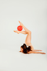 A girl gymnast in a beige leotard with a red ball does tricks on a white background. Front view