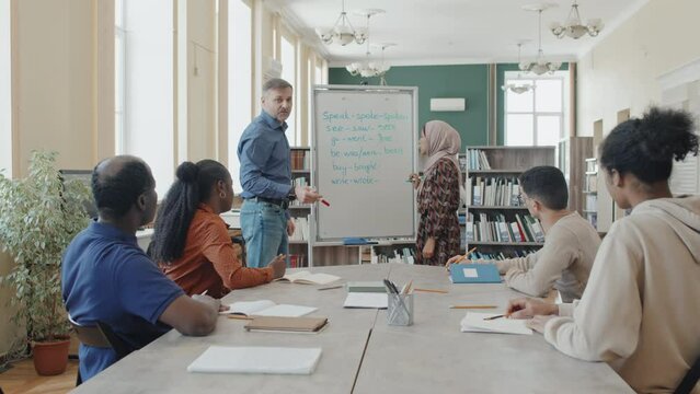 Group of ethnically diverse immigrants learning irregular verbs, female student wearing hijab writing past participle forms