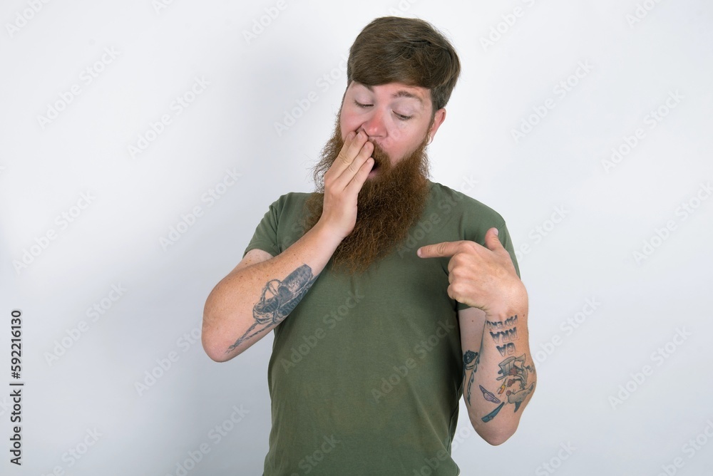 Wall mural Shocked Red haired man wearing green  T-shirt over white studio background look surprisedly down, indicates at blank  with fore finger, Scared model - Wall murals