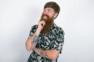 Red haired man wearing printed shirt over white studio background laughs happily keeps hand on chin expresses positive emotions smiles broadly has carefree expression