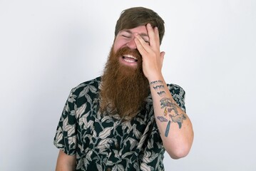 Red haired man wearing printed shirt over white studio background makes face palm and smiles...
