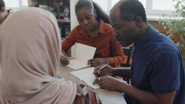 Senior Black man helping Muslim woman in hijab with exercise during language class explaining grammar rule to her