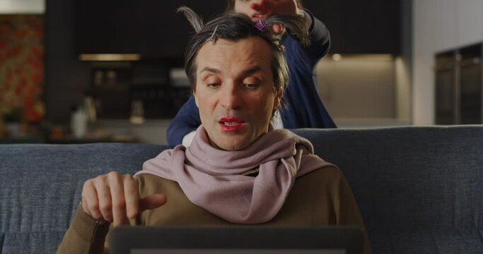 Funny Family Situation: Portrait of a Man Having a Video Call Conference with his Colleagues Using Laptop Computer at Home While his Daughter is Playing Makeup on him. Work from Home Concept