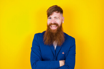 Coquettish red haired man wearing blue suit over yellow studio background smiling happily, blinking at camera in a playful manner, flirting with you.