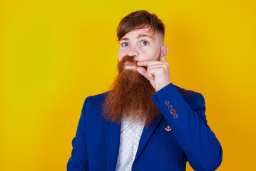 red haired man wearing blue suit over yellow studio background mouth and lips shut as zip with...