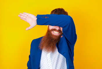 red haired man wearing blue suit over yellow studio background covering eyes with arm smiling...