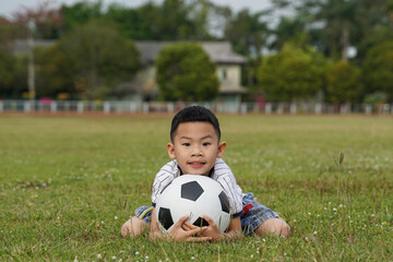 Naklejka premium Asian boy sits with his chin on a soccer ball in the grass, in the concept of outdoor activities, sports, playgrounds, leisure activities. Soft and selective focus. 