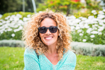 Attractive curly long blonde hair woman smiling at the camera with green park outdoors in...