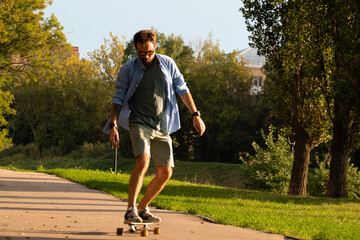 Front view of young guy riding on longboard along empty asphalt road between green trees
