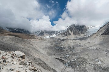Rocky relief surrounded by icy mountain peaks covered with fog and clouds under the blue sky