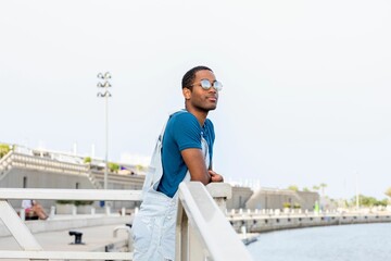 Young African American male in denim overalls and sunglasses posing on a pier