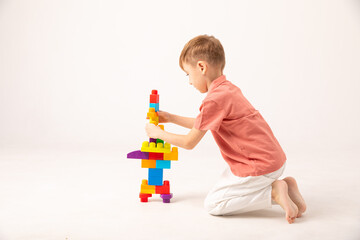 A happy child collects a tower of colored cubes. The preschooler sits down at the white background and assembles the construction set. The boy smiles, pleased with the result.