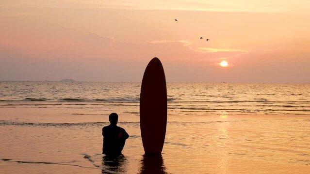 silhouette of surfer man sitting on the sea beach with long surf boards at sunset on tropical beach