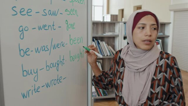 Muslim woman wearing hijab writing past participle forms of irregular verbs on whiteboard and explaining subject to students during English lesson