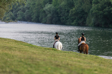 Riders, two young women riding beautiful horses down the calm river surrounded by the green grove