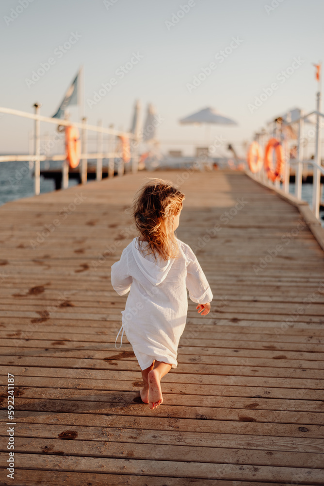 Wall mural a little girl in a white dress and long hair runs barefoot along a wooden pier towards the ocean at 