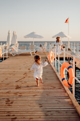 A little girl in a white dress and with long hair runs barefoot along a wooden pier towards the ocean to her mother. Beautiful light and wind in the hair