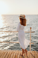 A young girl in a white dress and long hair, in sunglasses and a straw hat, stands on the pier against the backdrop of the ocean at sunset