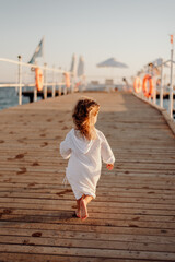 Fototapeta premium A little girl in a white dress and long hair runs barefoot along a wooden pier towards the ocean at sunset. Beautiful light and wind in the hair