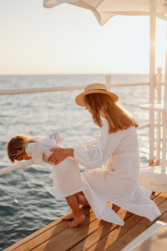 A young girl, mother, holds her little daughter by the waist. The daughter looks into the water. Girls in white dresses on the background of the evening ocean